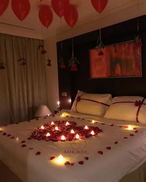 اتاق عروسی که روتختی آن با گلبرگ های رز قرمز و شمع های روشن به شکل قلب و سقف آن با بادکنک های هلیومی قرمز که گل رز از آن ها آویزان شده است، تزیین شده است